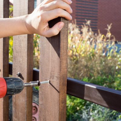 Close up of hand screewing wood plank to metal construction. Building a wooden fence with a drill and screw.