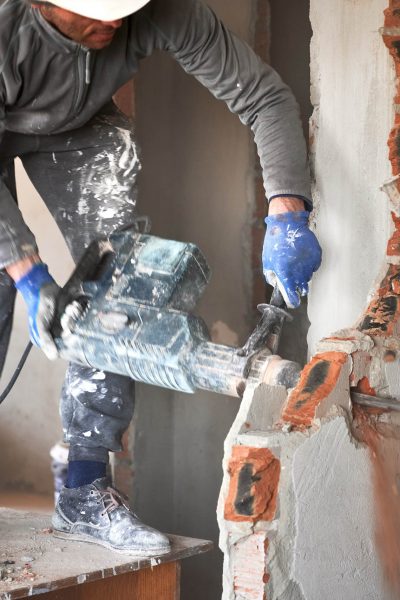 Close up of male builder in workwear drilling wall with hammer drill. Man worker using drill breaker while destroying wall in apartment under renovation. Demolition work and home renovation concept.